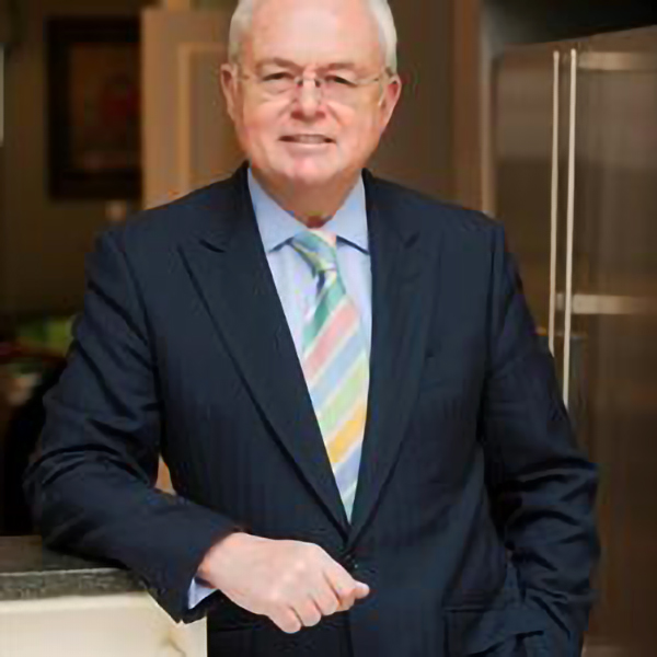 Headshot of Martyn Lewis, Demelza celebrity ambassador, smiling at the camera in a navy suit, pale blue shirt, and frameless glasses.