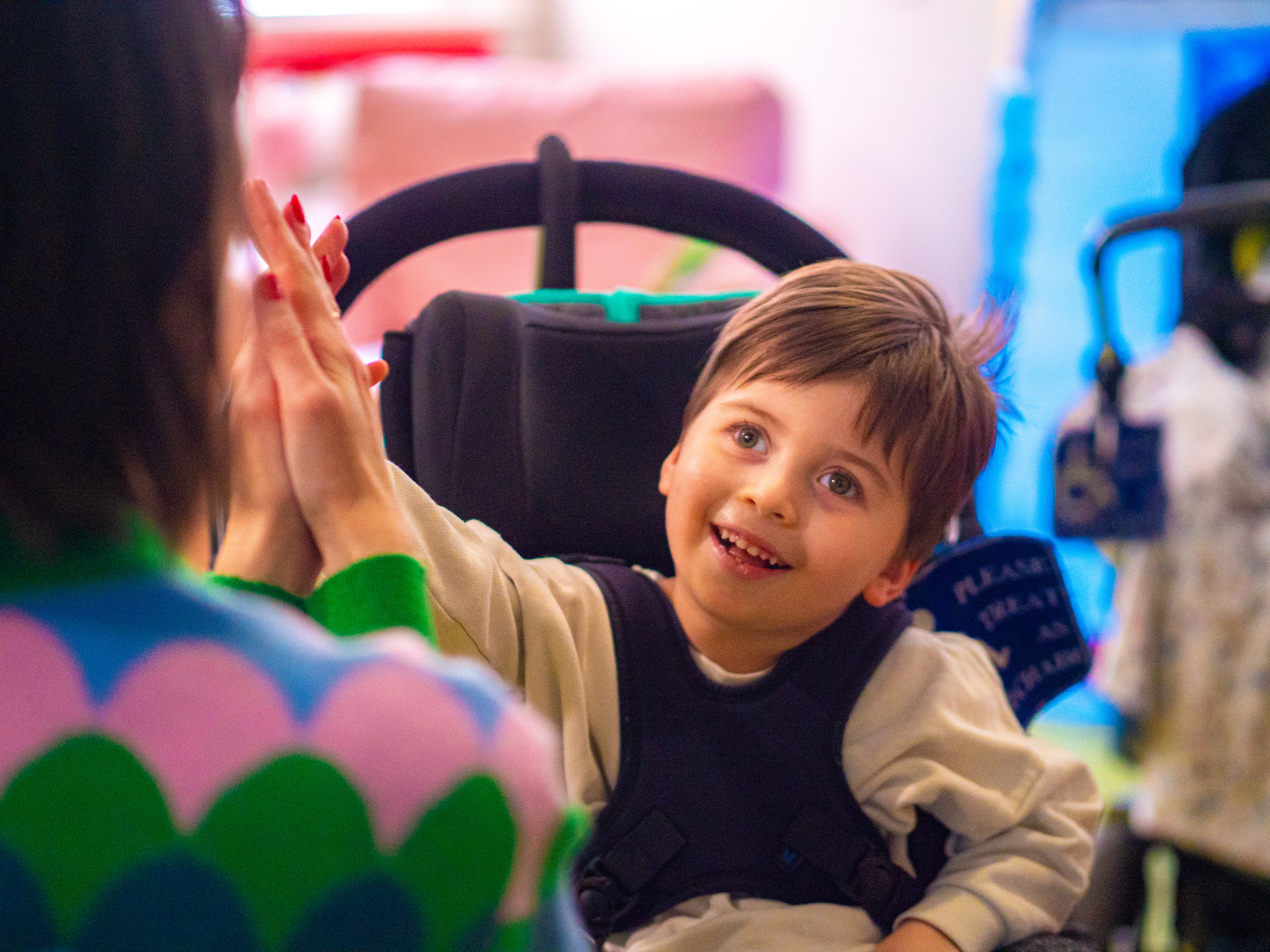 A young boy with brown hair in a wheelchair looking towards his mum, a woman with black hair and red nails, smiling and putting his hand out.