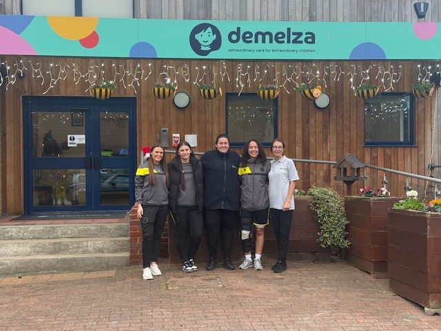 Five people stand in front of a building with a 'demelza extraordinary care for extraordinary children' sign, decorated with bee-themed ornaments and string lights.