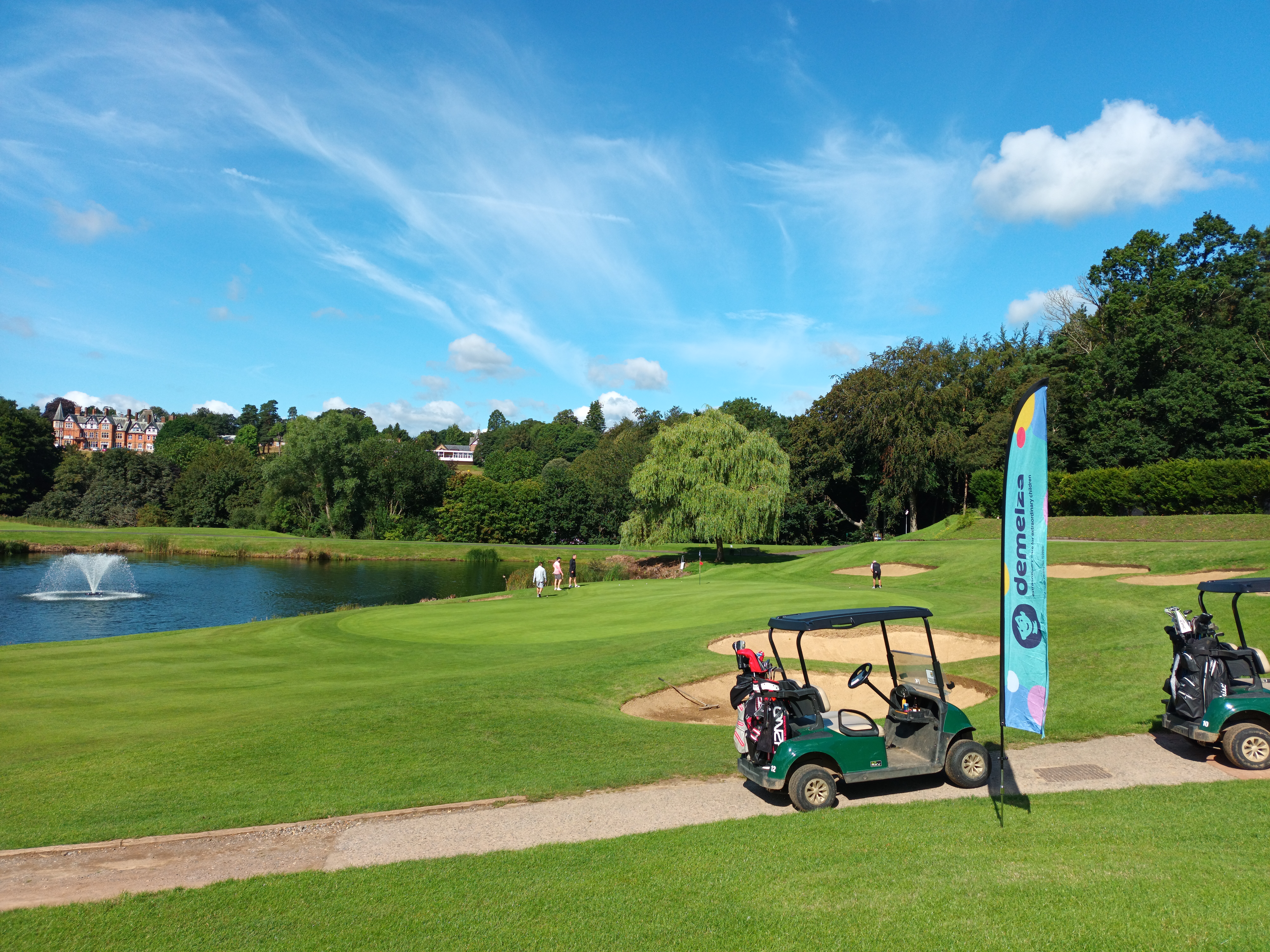 Scenic golf course with two golf carts on a path, a "Demelza" flag nearby, and a pond with a fountain under a clear blue sky with surrounding greenery.