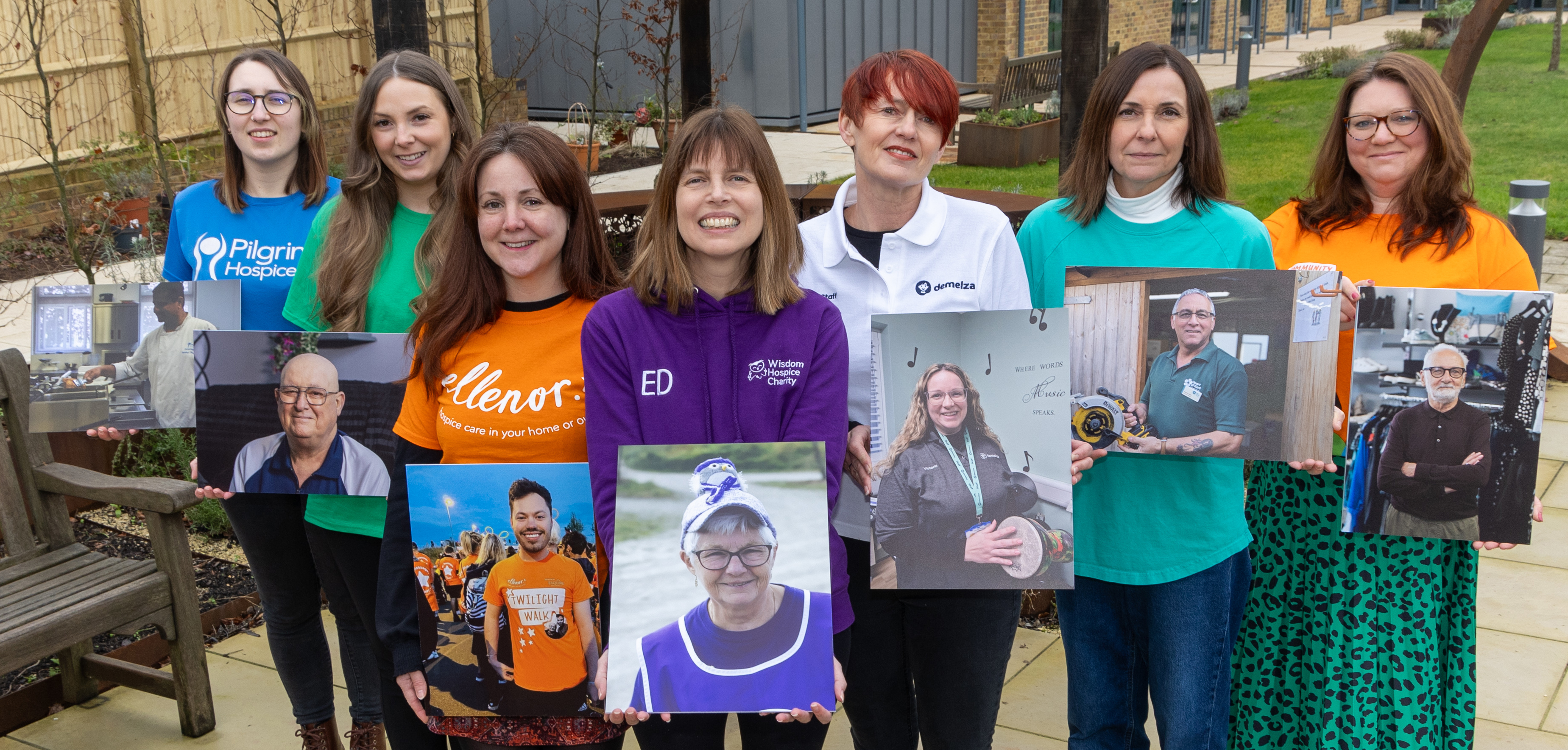 Group of seven people outdoors holding photos of others; some wear shirts with "Demelza", "Pilgrims Hospice" and "ellenor" logos.
