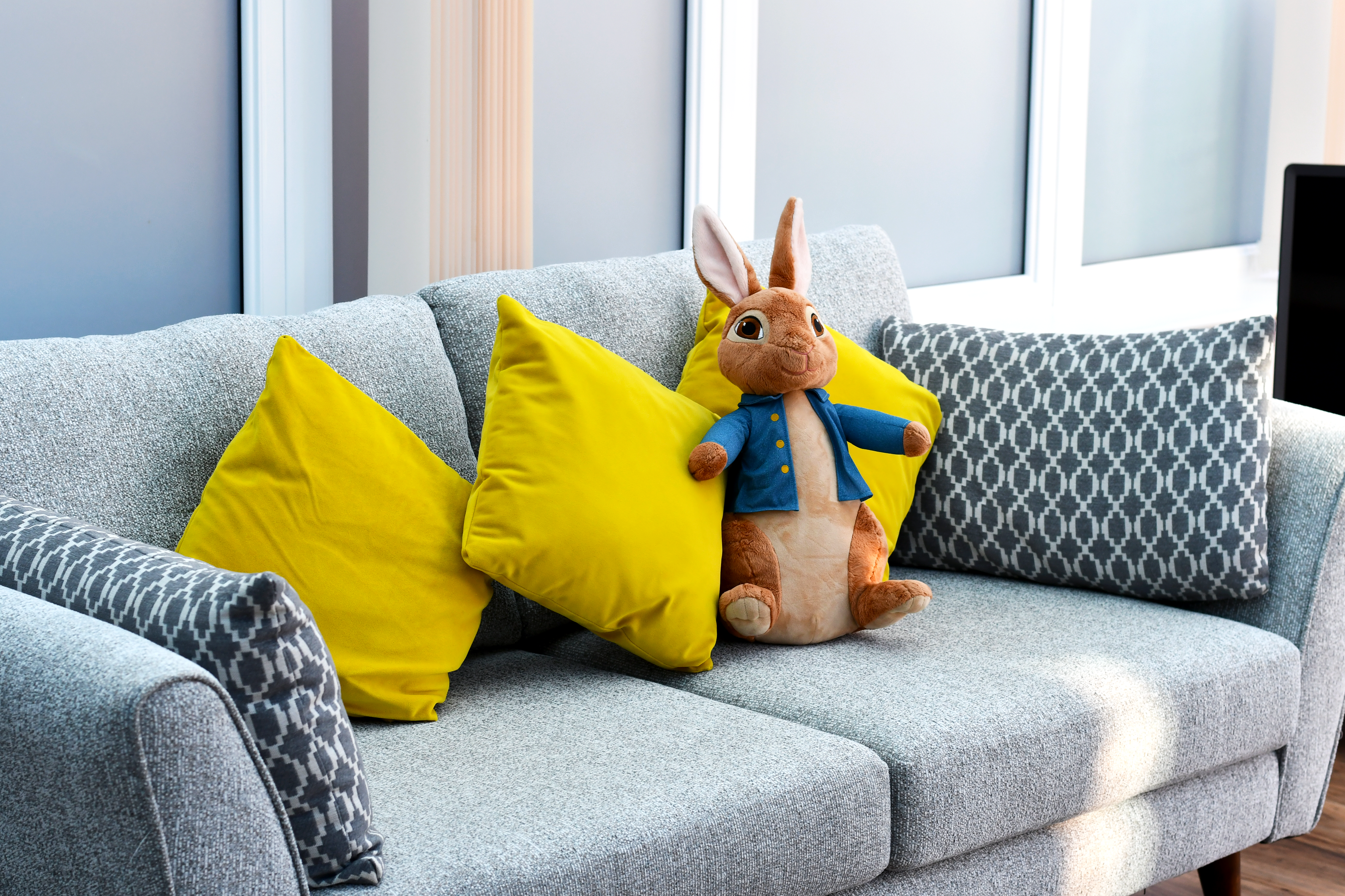 A grey sofa with two yellow pillows and two patterned grey pillows. A stuffed rabbit toy wearing a blue jacket sits in the centre of the sofa. Large windows with white frames are visible in the background.