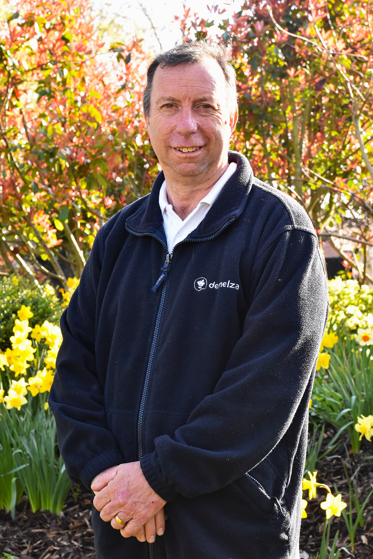 A man with brown hair wearing a blue Demelza jacket outside in the tranquility garden smiling at the camera