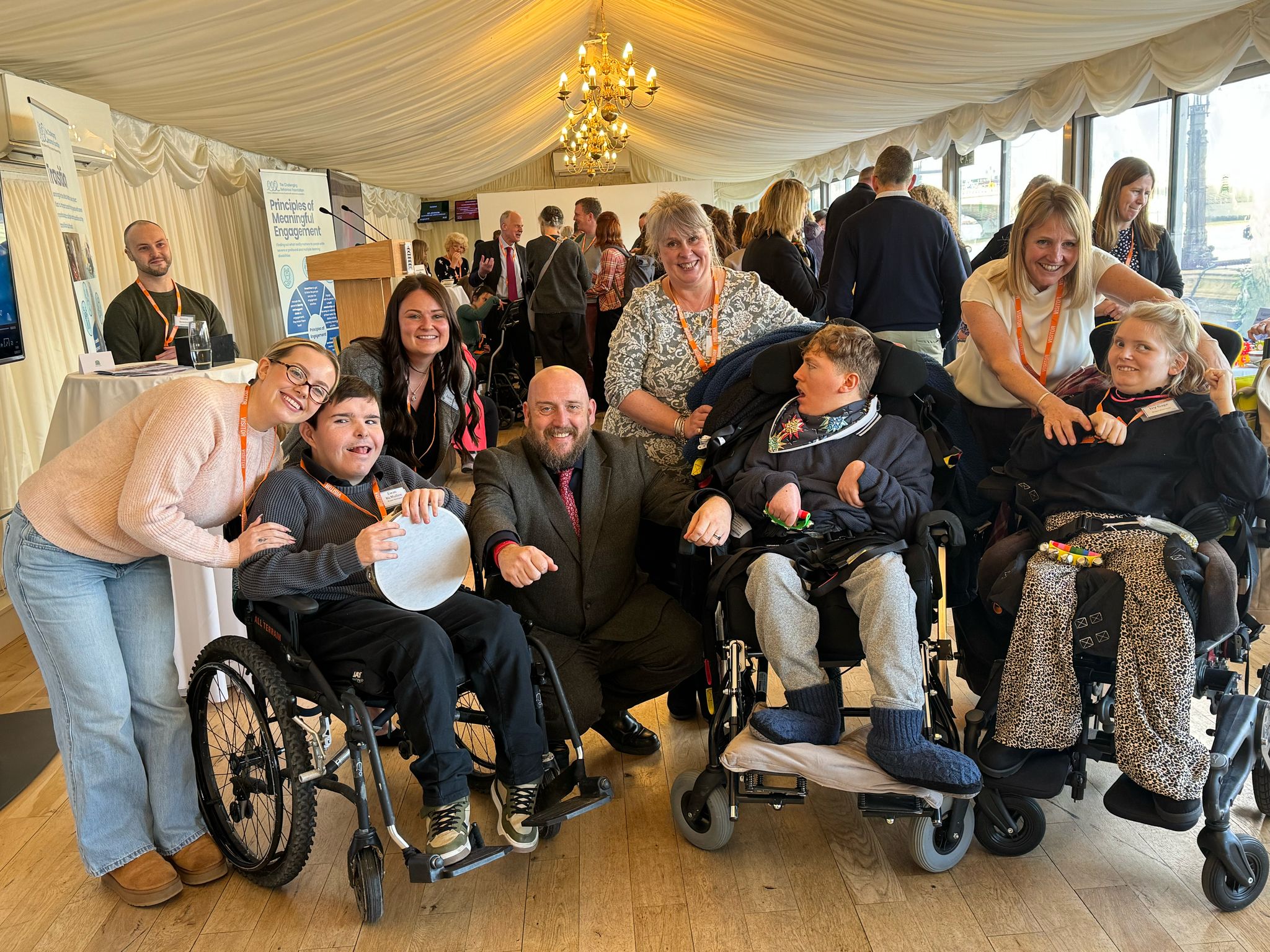 Group of people, including wheelchair users, gathered in a large room with chandeliers, draped ceilings, and a podium in the background. Some are seated, others standing.