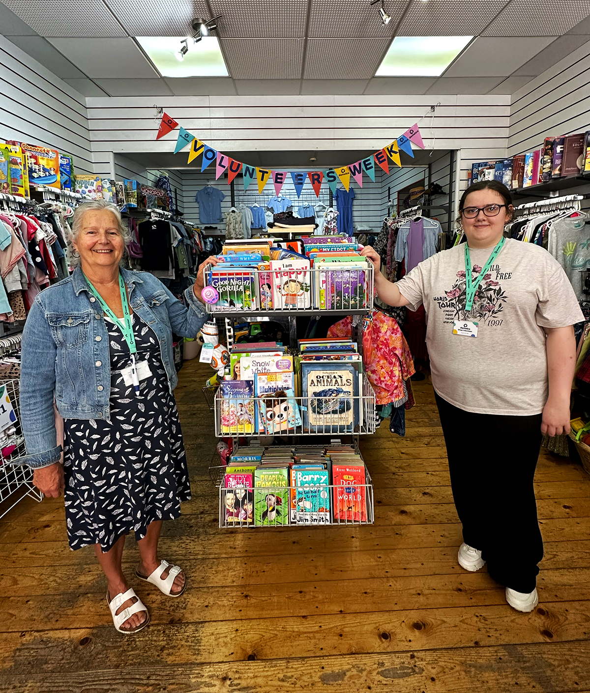 Volunteers Abigail and Maggie smile proudly in Demelza’s Sheerness Shop during Volunteers Week, standing by a colourful book display