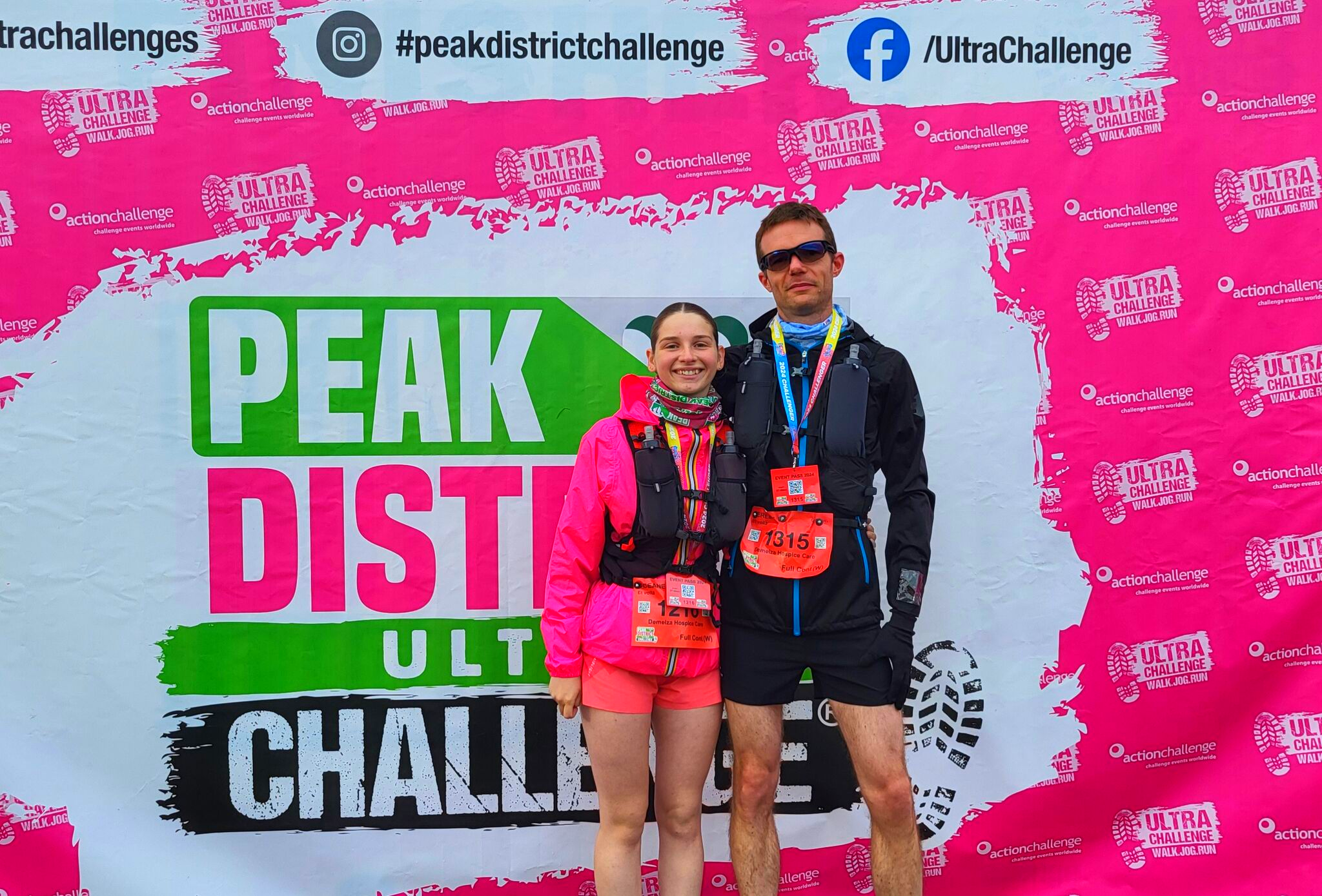 Two people in athletic gear with medals pose in front of a "PEAK DISTRICT ULTRA CHALLENGE" backdrop featuring hashtags and social media handles.