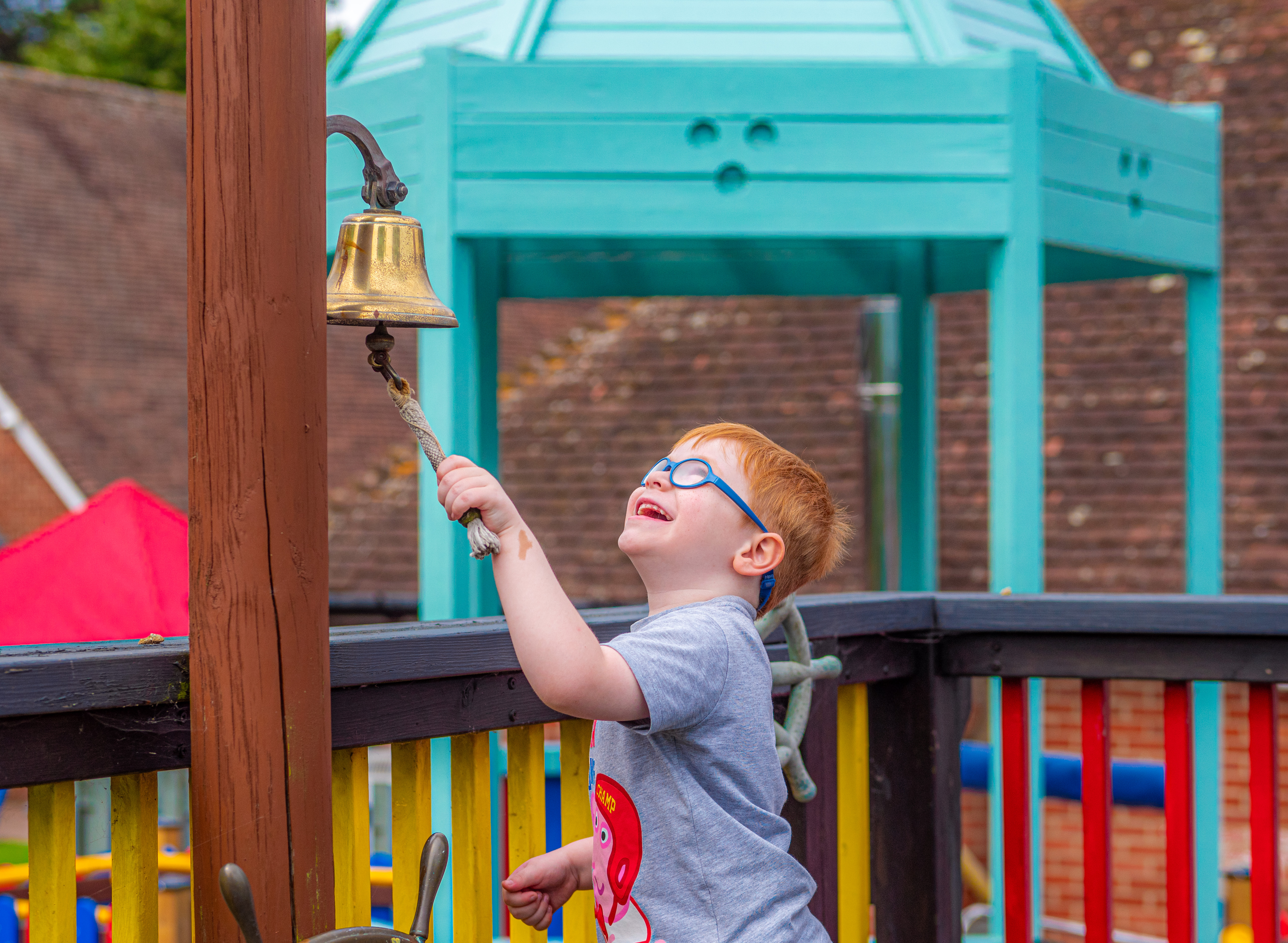 A young boy with glasses ringing a bell in the playground at Demelza Kent