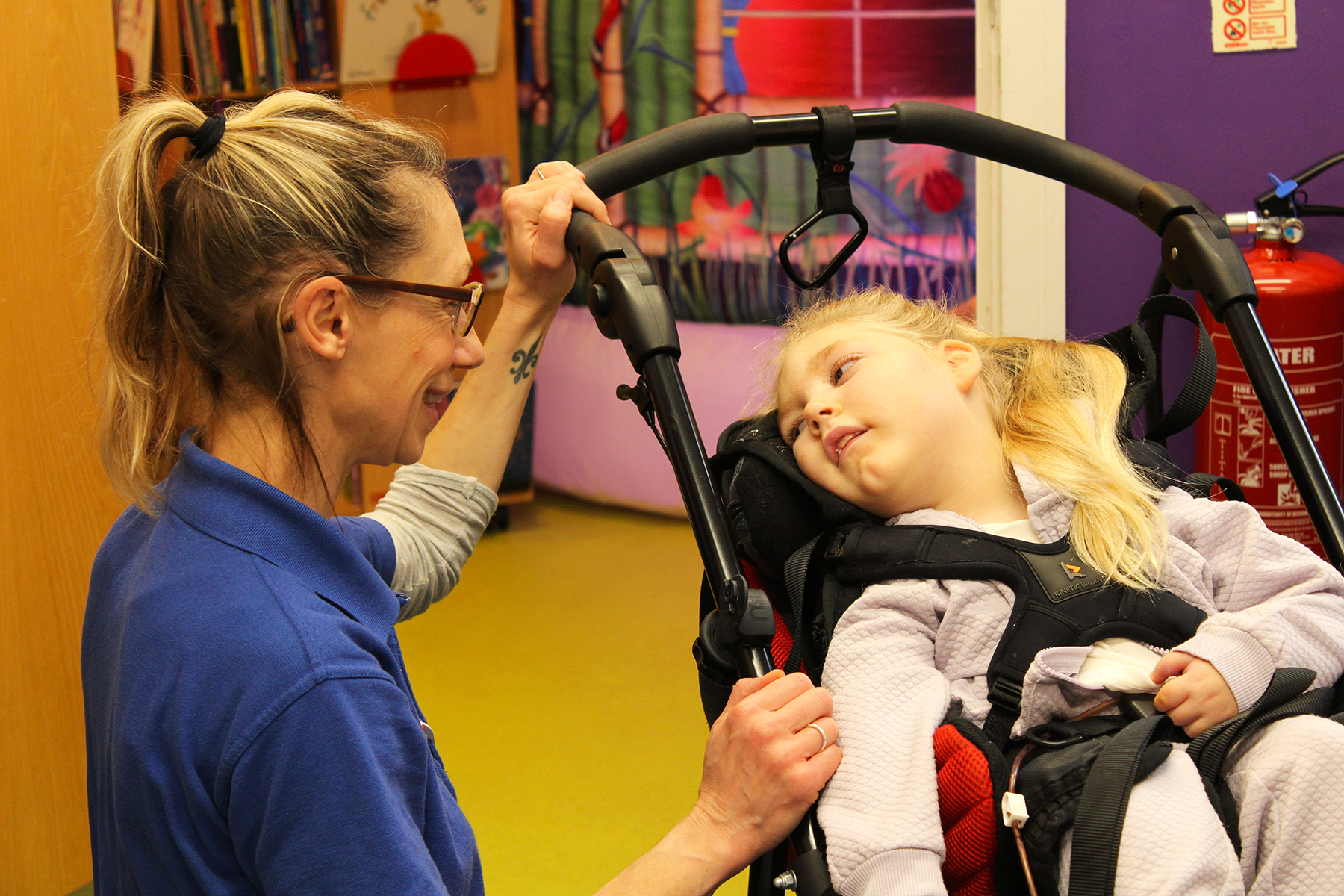 A nurse in a blue Demelza shirt, blonde hair and glasses, smiling towards a young girl with blonde hair in a wheelchair