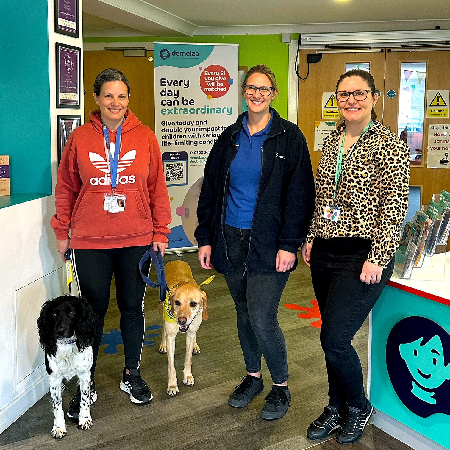 Three people stand indoors with two dogs. Behind them is a banner reading: 'Every day can be extraordinary. Give today and double your impact for children with serious conditions.'