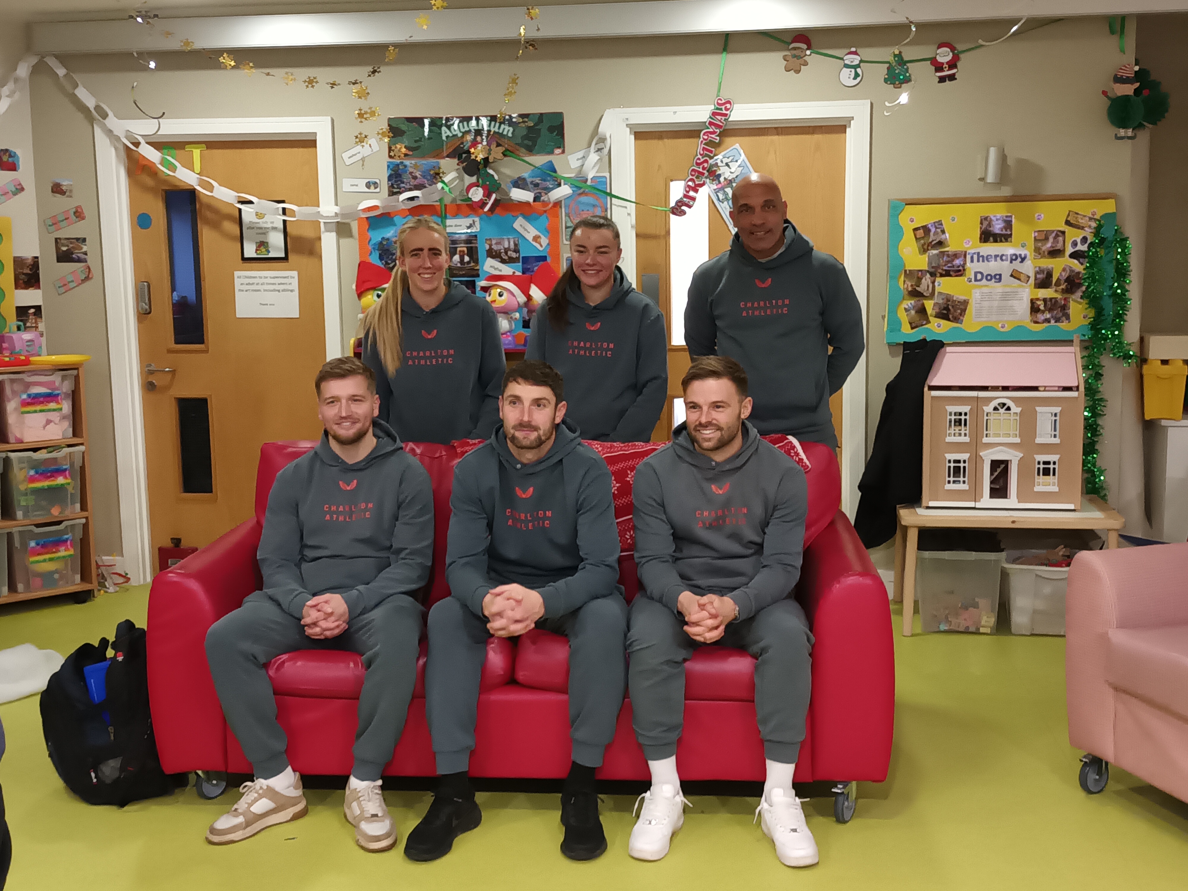Six people in matching grey football training outfits sit and stand around a red couch in a decorated room with children's toys and artwork.