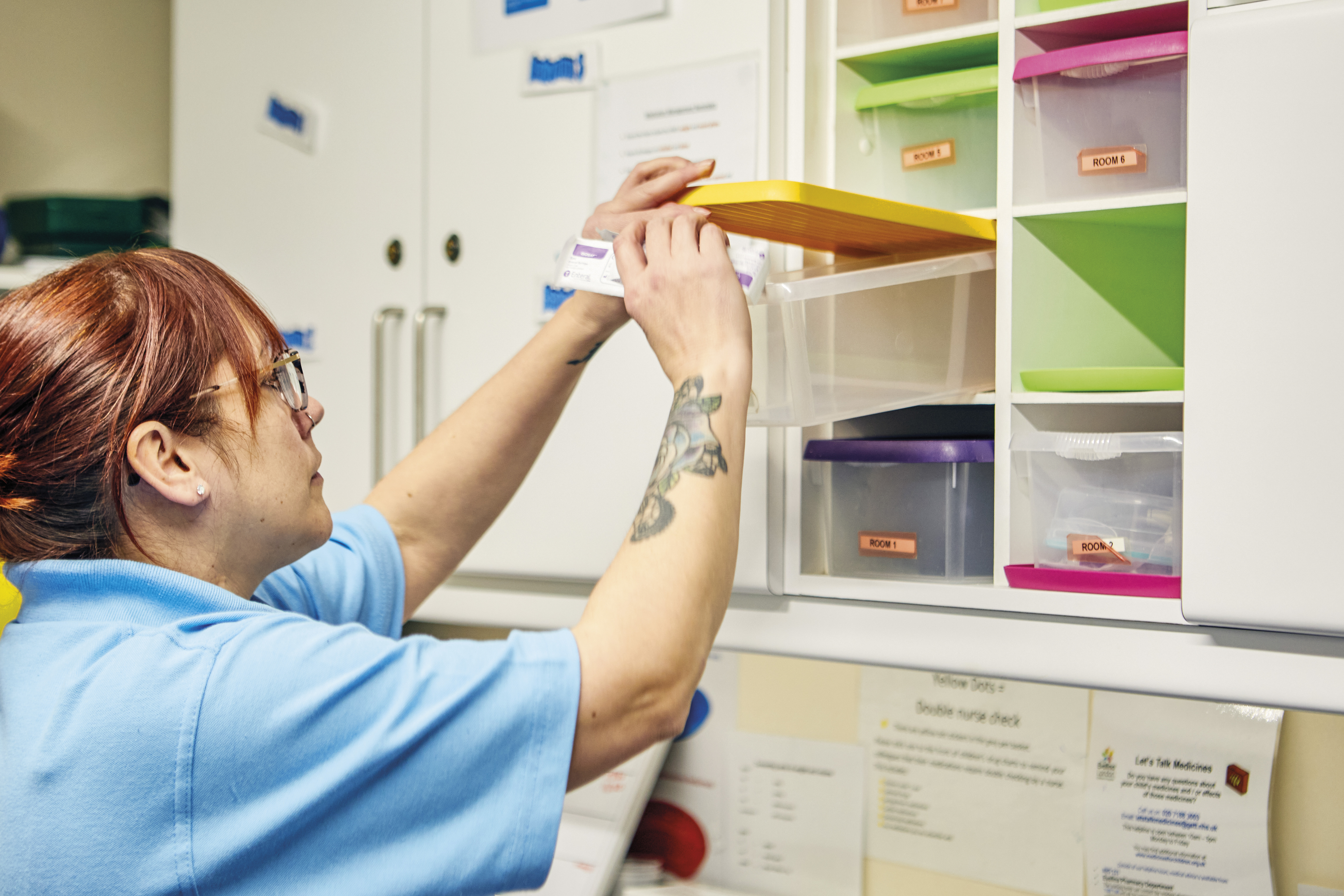 Nurse Becky in blue uniform reaches into a yellow-lidded bin labelled by Room number to sort medications in a storage unit.