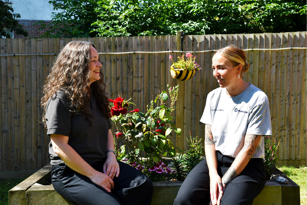 Two female Care Team staff members sitting outdoors in the sunny East Sussex gardens, surrounded by blooming flowers.