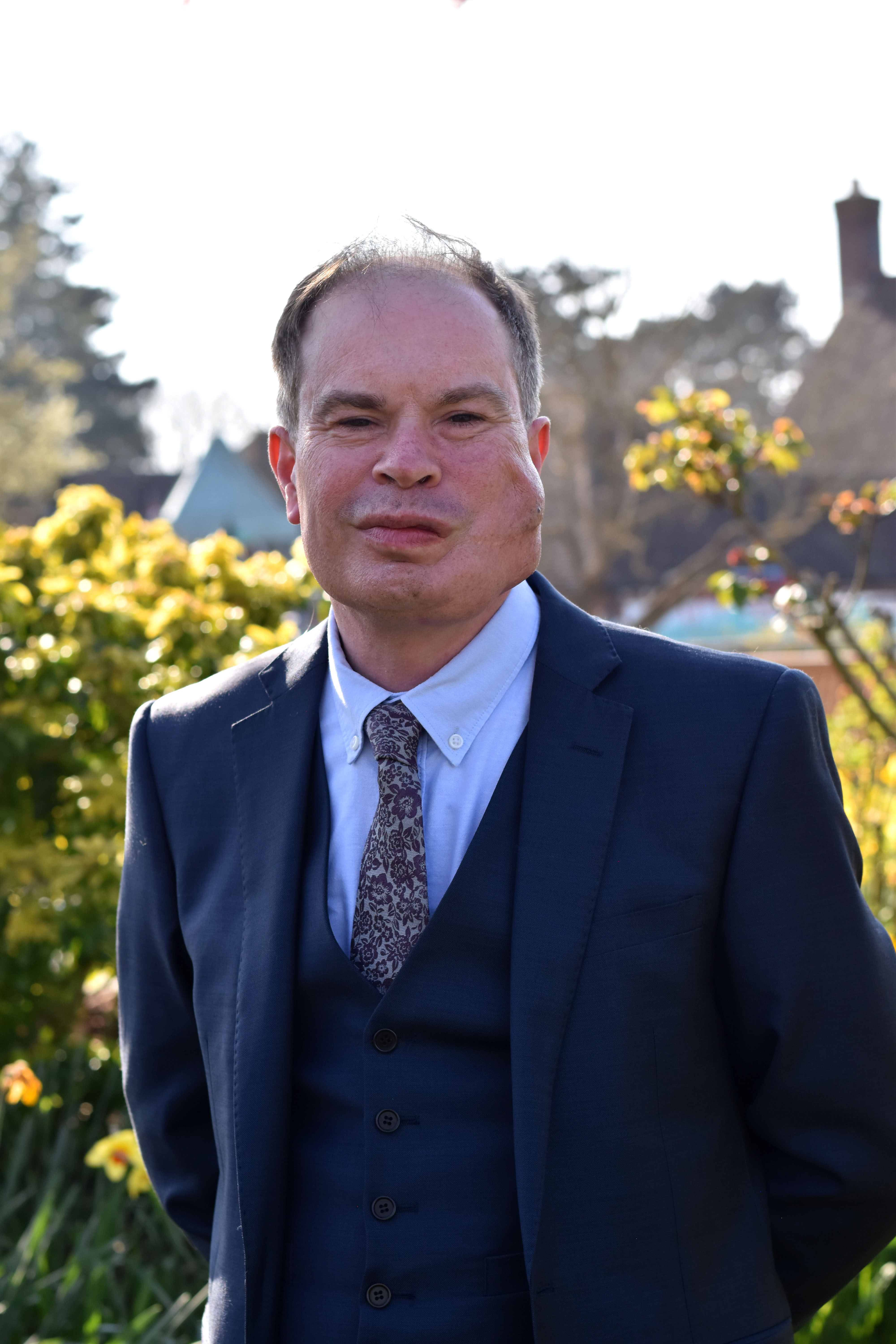 Chair of Safeguarding Assurance Committee Dr Richard Patey stands in sunny gardens wearing a smart navy blue three-piece suit and tie.
