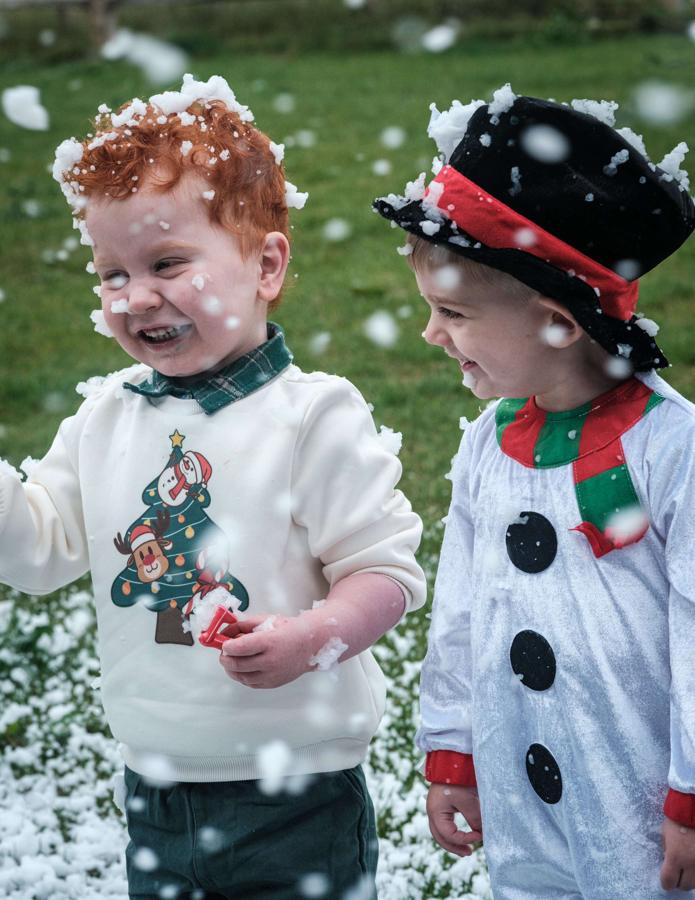 Two young boys smiling wearing Christmas jumpers whilst snow falls around them