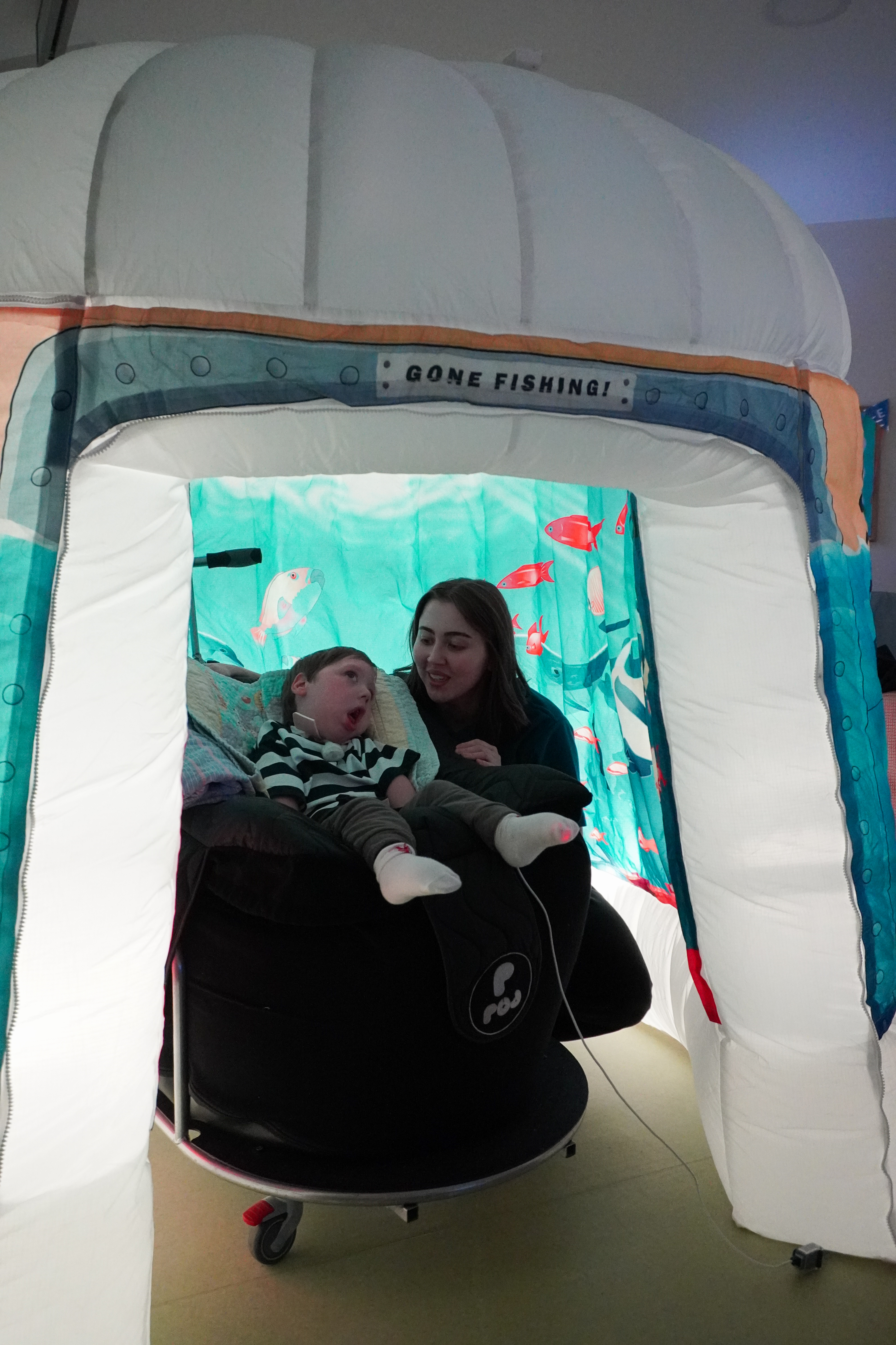 A nurse and a child cared for by Demelza sit inside an inflatable structure with a 'Gone Fishing!' sign. Background shows an underwater scene with fish and aquatic plants.