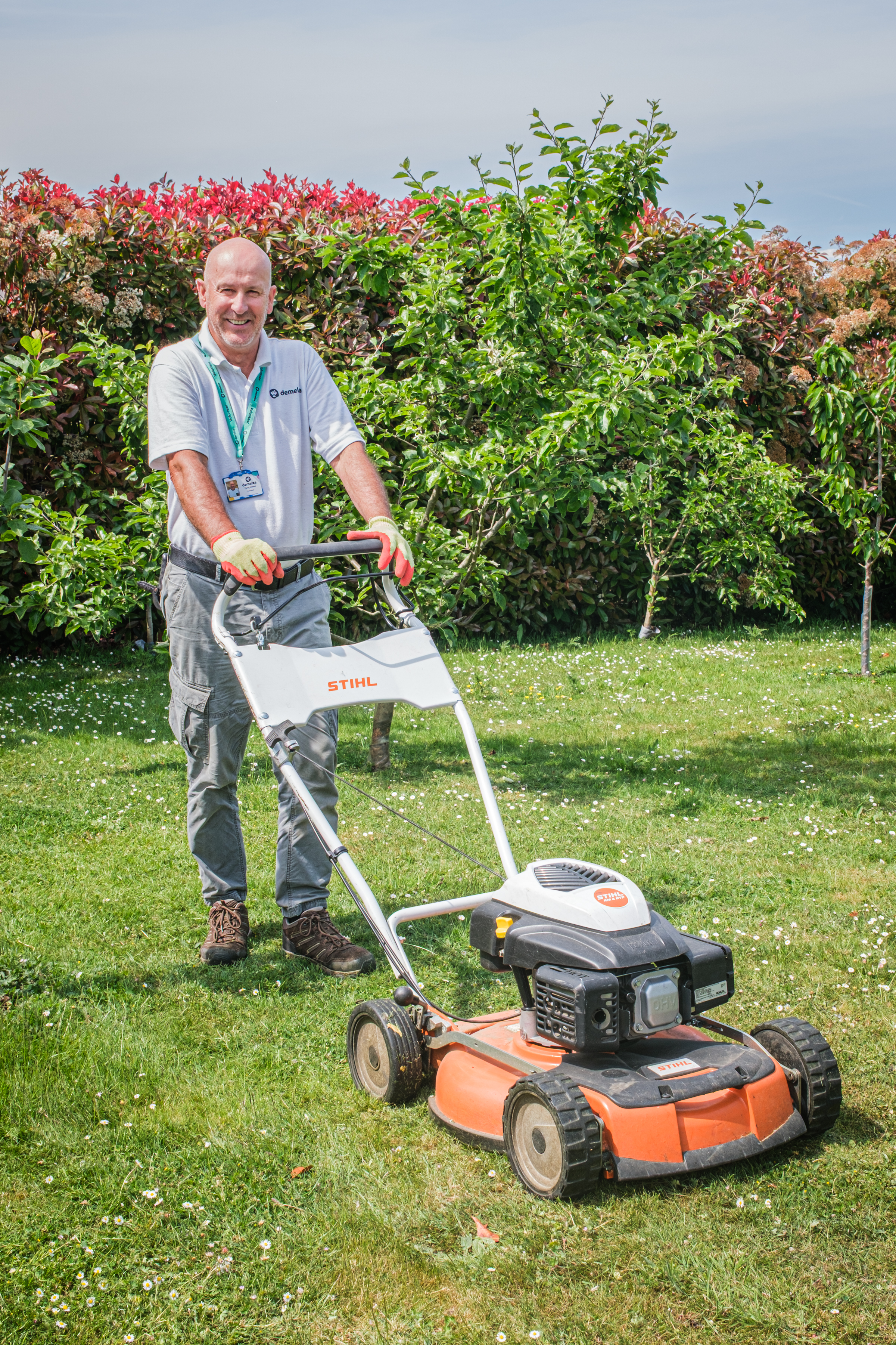 Smiling male volunteer pushes a lawn mower across a sunlit garden, trimming grass under clear blue skies.