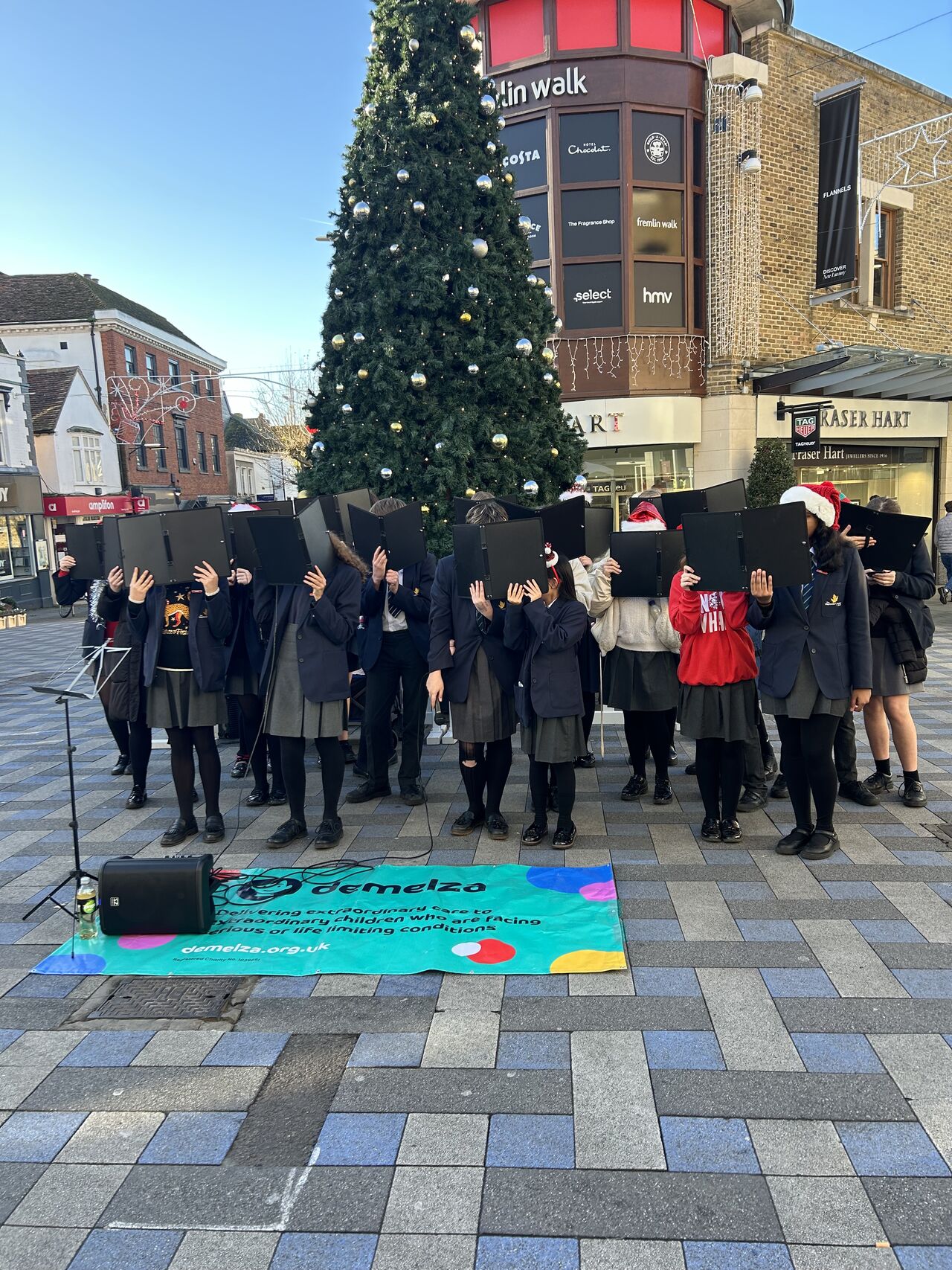 Group singing in front of a large Christmas tree in a town square. Some wear Santa hats. 'Demelza' charity banner on the ground.
