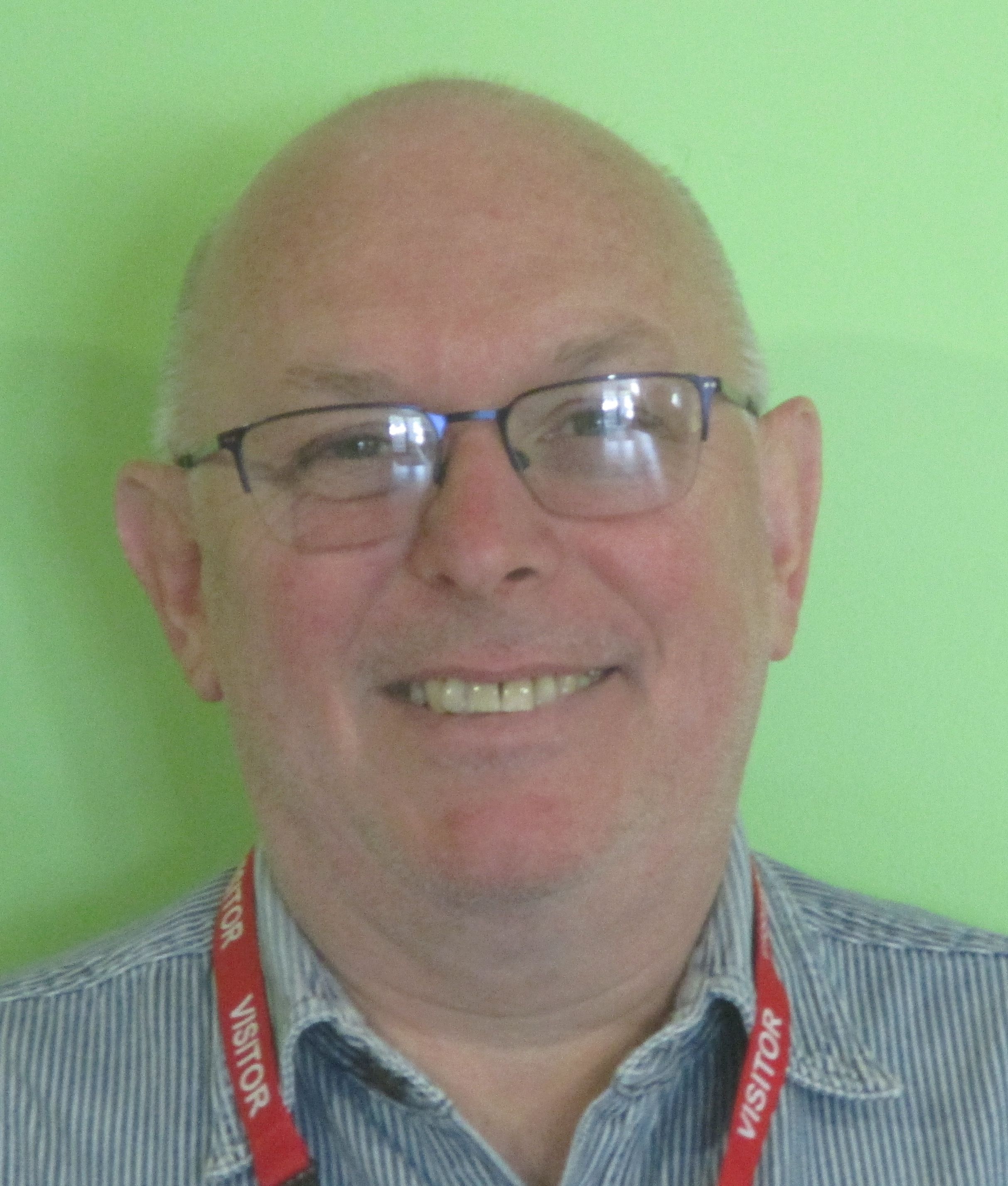 Person wearing a blue and white striped shirt and red "VISITOR" lanyard, standing in front of a green background.