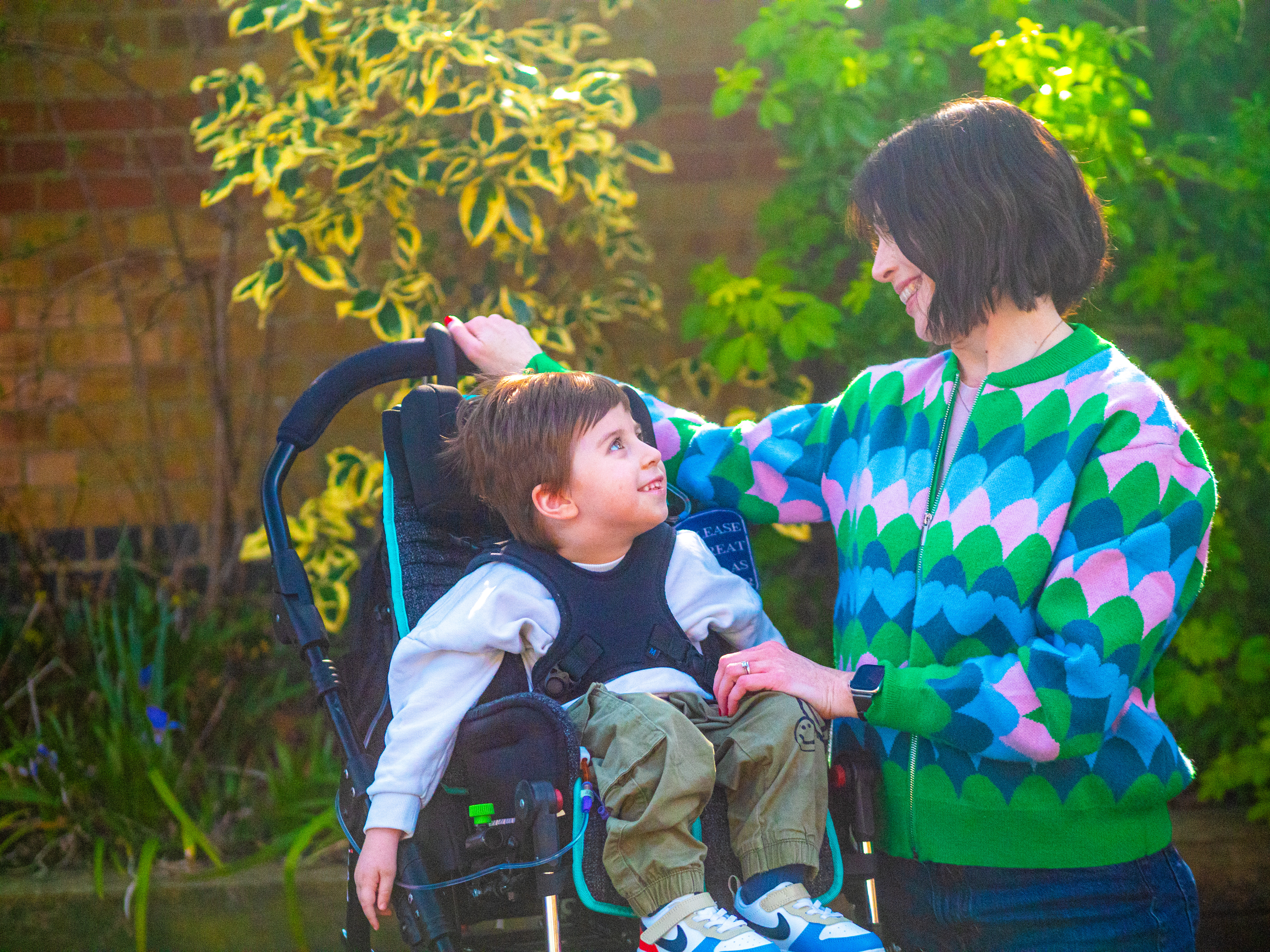A woman with a green, blue and pink patterned cardigan looking at her son who has a white shirt and is in a wheelchair smiling