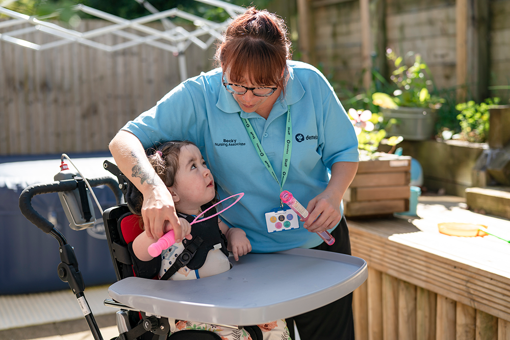 A woman in a light blue shirt blowing into a bubble wand with a young girl sitting in an arm chair