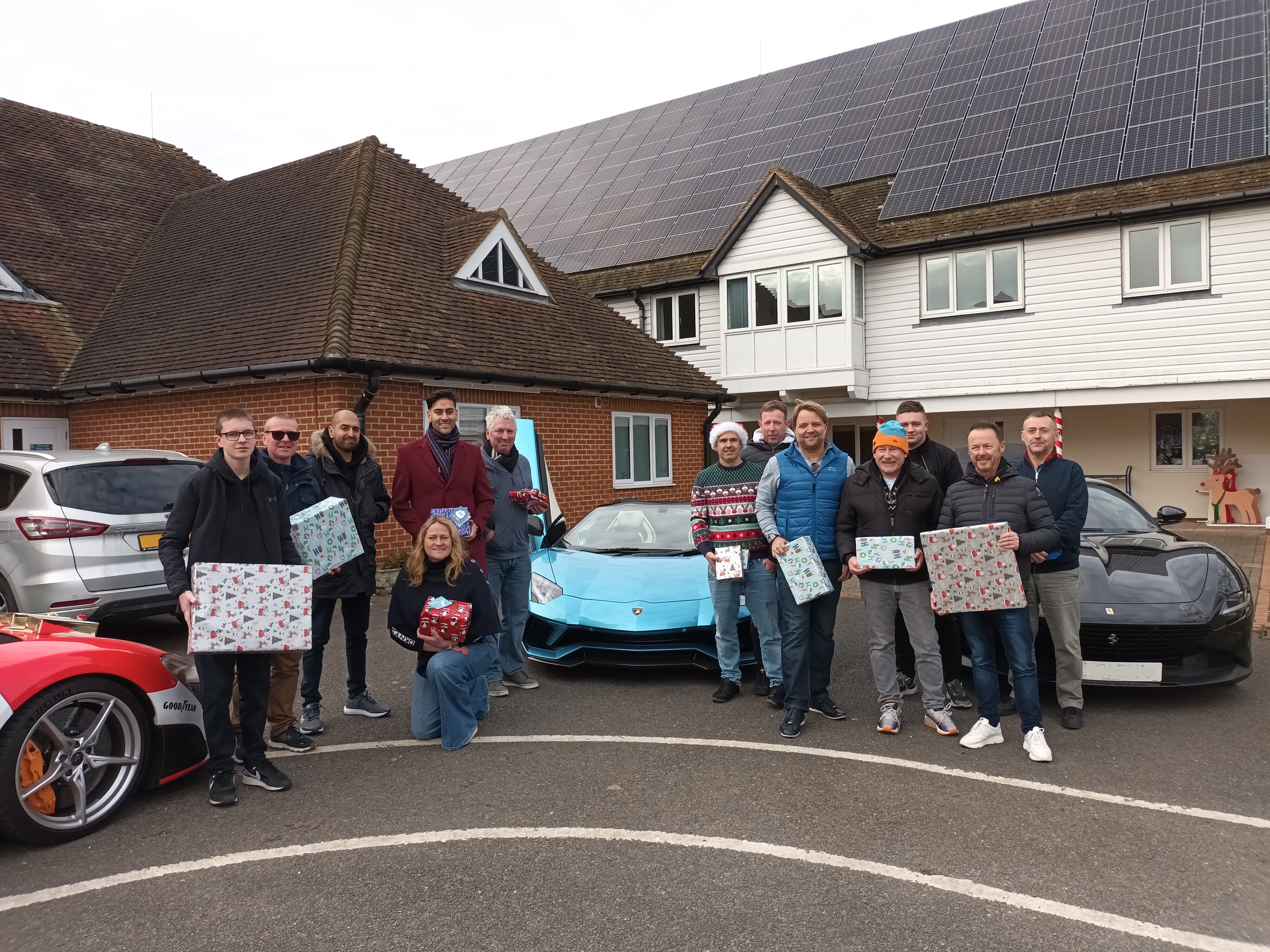 Group of people holding wrapped gifts in front of a Demelza building; several luxury cars are parked nearby.